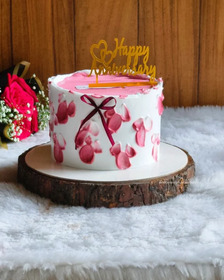 White anniversary cake with pink floral petals, red ribbon, and a 'Happy Anniversary' topper.
