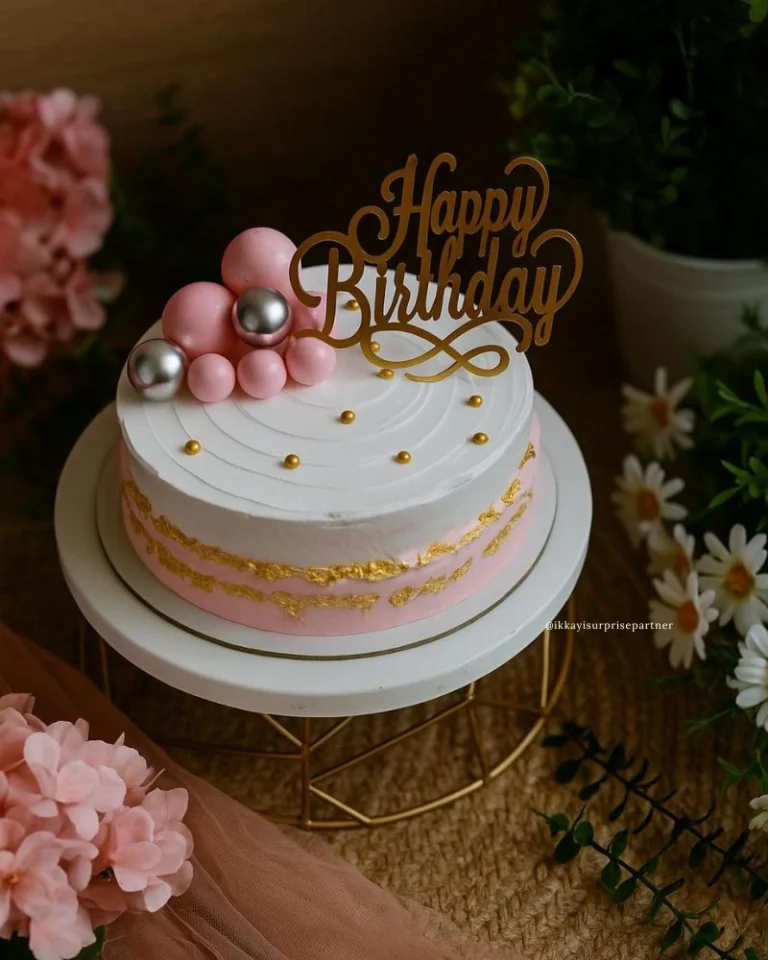 Pink and white birthday cake with gold foil, pink and silver spheres, and 'Happy Birthday' topper.