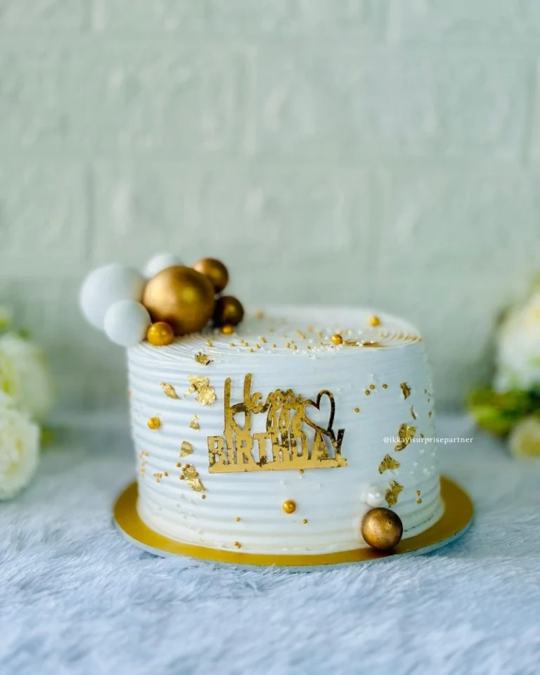 White birthday cake with gold leaf flakes, metallic gold spheres, and a 'Happy Birthday' topper.